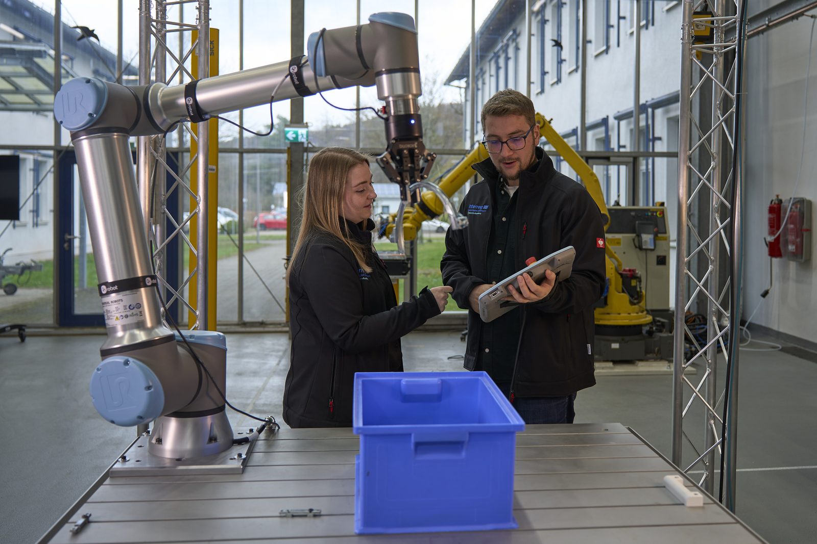 Eine Frau und ein Mann stehen in einer Technikumshalle an einem Tisch mit einem kollaborativen Roboterarm; der Mann hält ein Tablet, auf dem Tisch steht eine blaue Transportkiste.