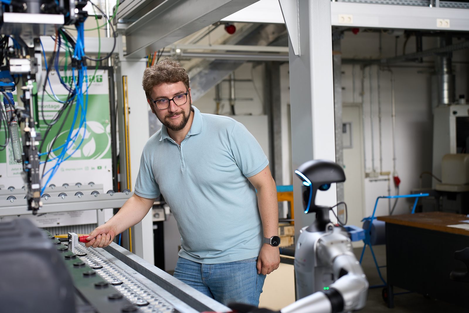 Ein Mann bedient eine Montagelinie; von rechts greift ein kollaborativer Roboterarm in den Arbeitsbereich.